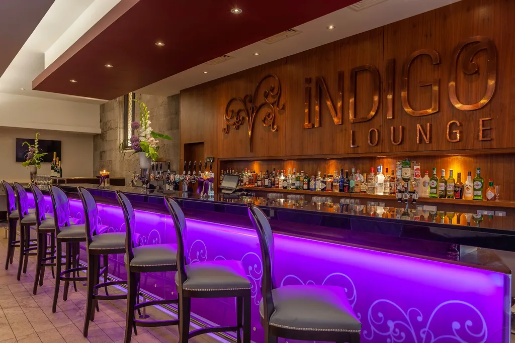 Indigo Lounge's purple-lit bar with grey high-backed stools beneath a bronze Indigo Lounge sign on a walnut wall