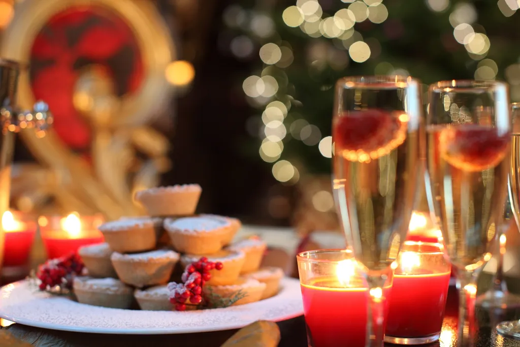 Mince pies dusted with icing sugar beside glasses of sparkling wine with raspberries and red candles