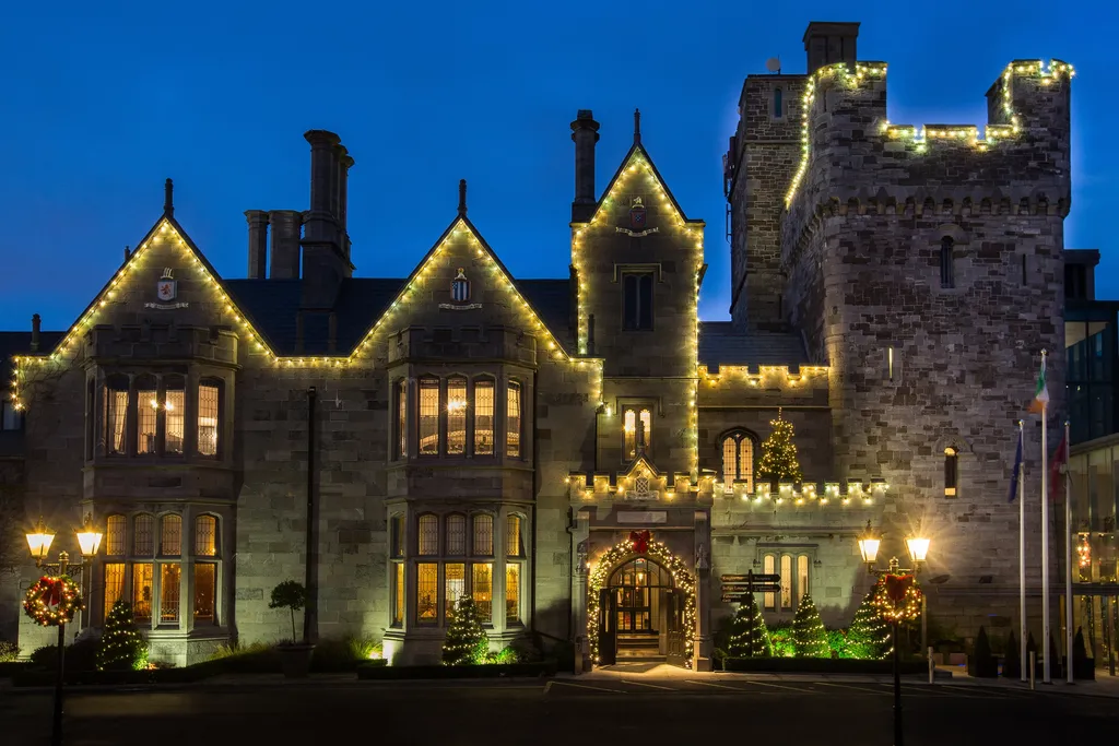 The castle’s gabled facade and tower at twilight, every gable and battlement traced in fairy lights, with lit trees and lanterns at the entrance