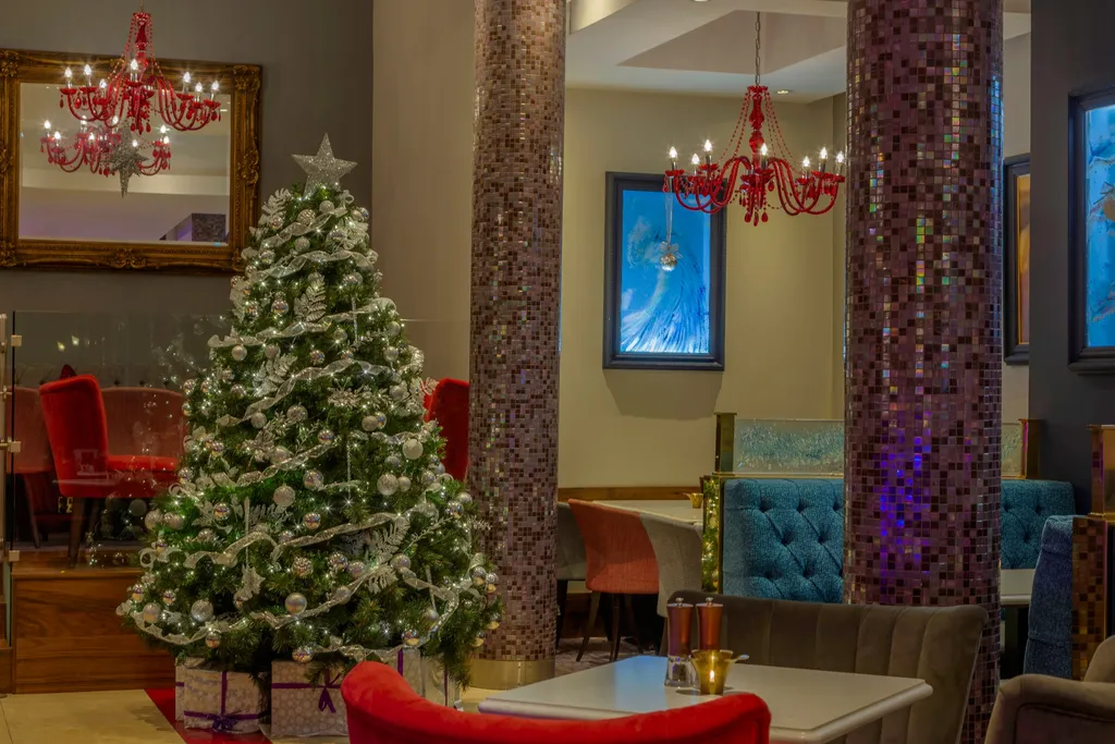 A white-and-silver Christmas tree among red chairs and teal booths in Indigo Lounge, beneath red glass chandeliers