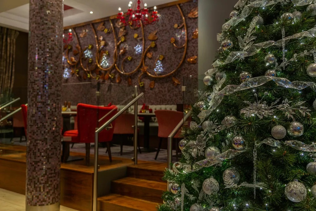 A Christmas tree with silver ribbon and baubles beside a mosaic pillar in Indigo Lounge, with red chairs on the raised seating area