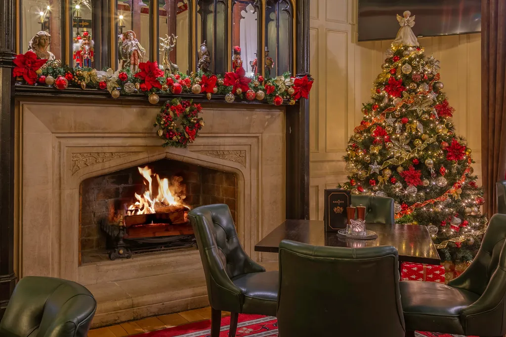 The Knights Bar’s stone fireplace with a log fire, its carved Gothic overmantel decked in poinsettias and garland, beside a red-and-gold Christmas tree and green leather chairs