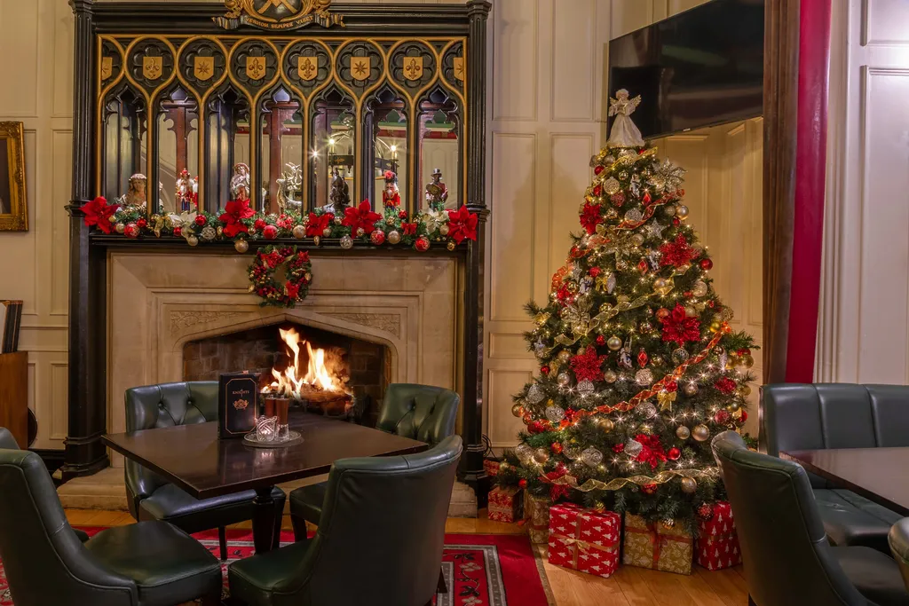 The Knights Bar’s garlanded fireplace beneath its Gothic carved overmantel, with a red-and-gold Christmas tree and wrapped presents