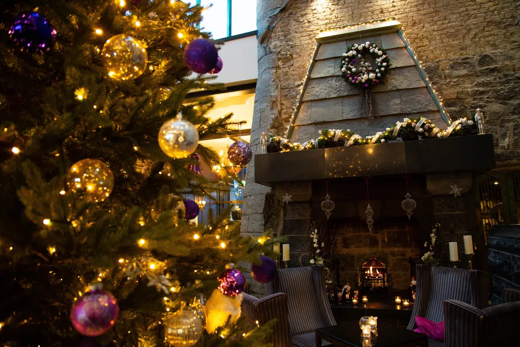Gold and purple baubles on the lobby Christmas tree, with the garlanded stone fireplace and a wreath beyond