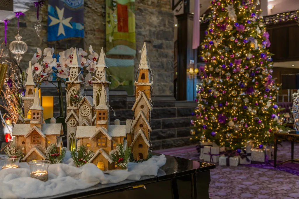 A miniature snowy village of wooden churches with white orchids, before a lit Christmas tree and heraldic banners in the lobby
