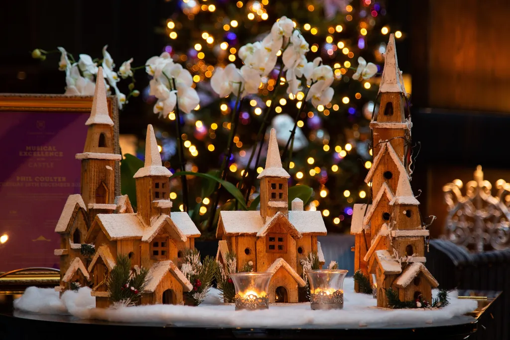 A wooden Christmas village of snow-dusted churches with white orchids and tealights, before a glowing tree