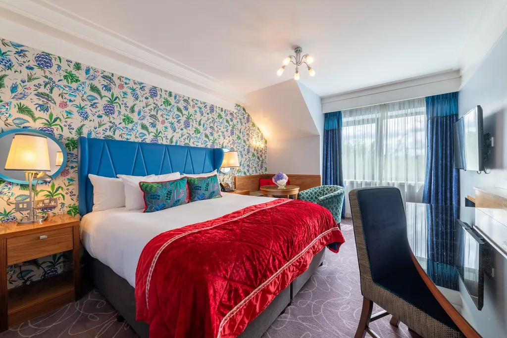 Deluxe Room with a blue headboard, red throw, desk chair and tall windows with blue curtains