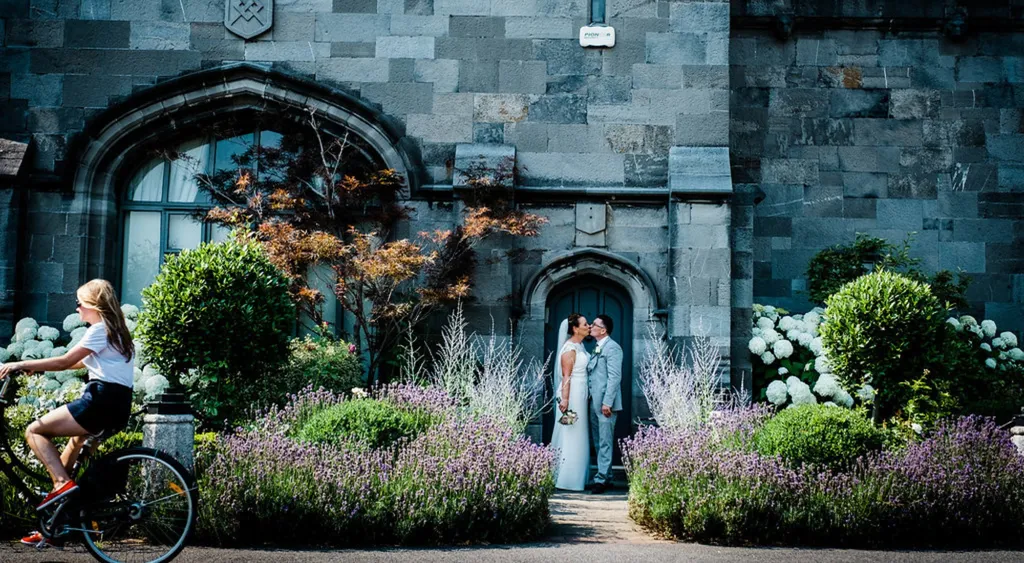 A bride and groom kissing in an arched stone doorway framed by lavender as a cyclist rides past