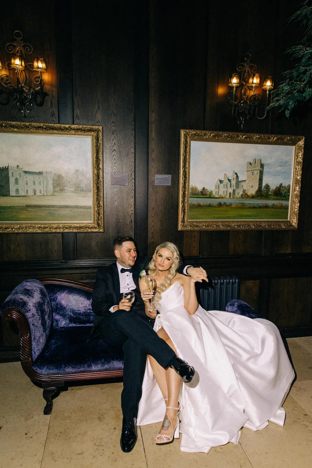 A bride and groom laughing together on a purple velvet chaise in a wood-panelled room lit by wall sconces