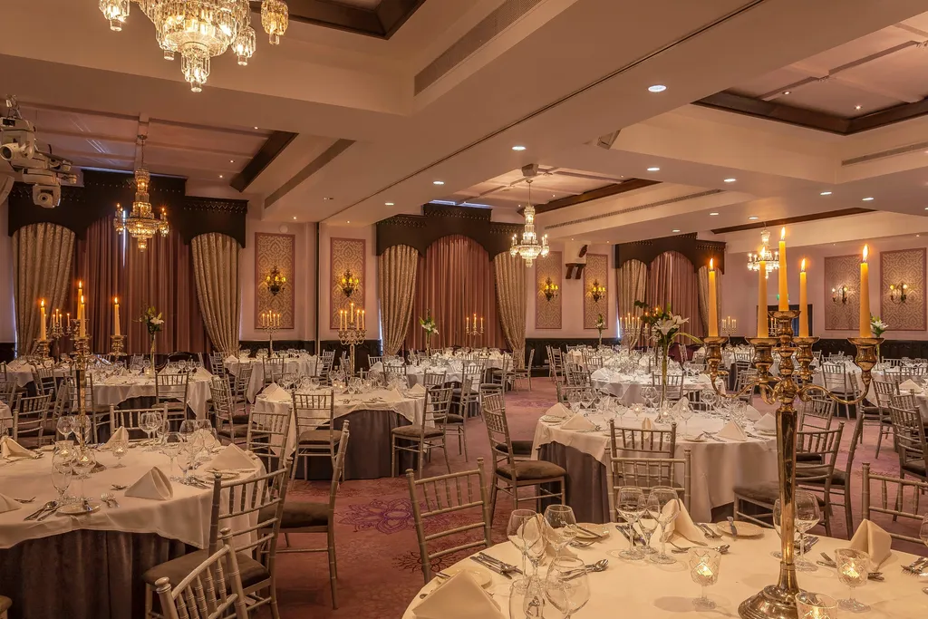 A warmly lit ballroom laid with round tables, gold chairs and candelabra centrepieces beneath chandeliers